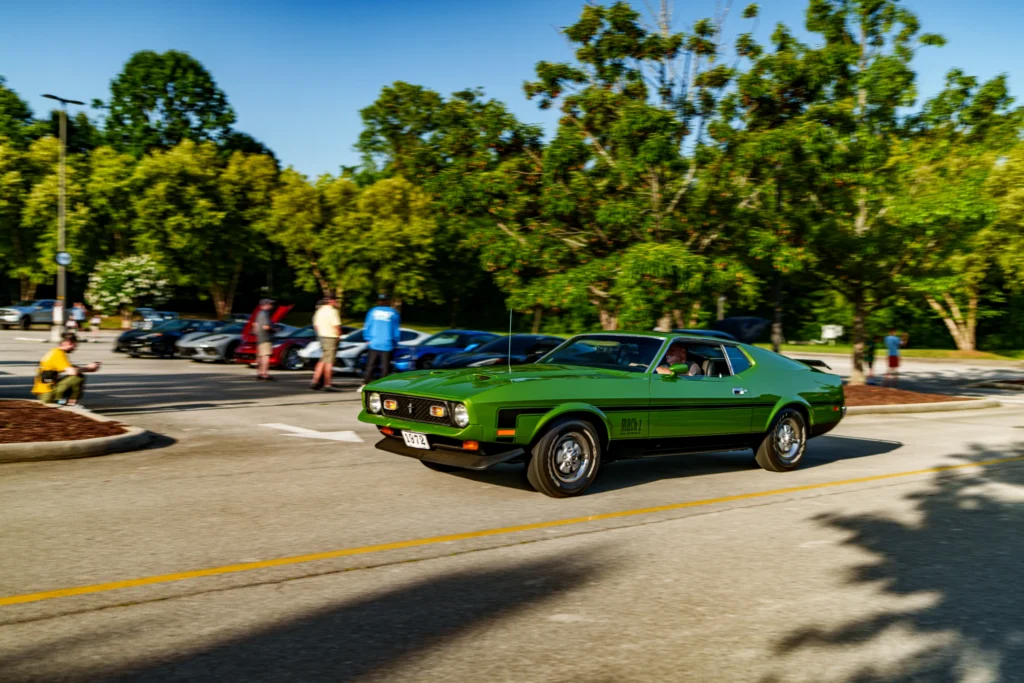 Green Ford Mustang Mach 1 driving on a street with other cars and people in the background at an outdoor event