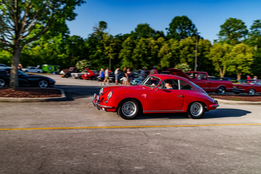 Red Porsche 356 coupe driving in parking lot at a car meet with other vehicles and people in background