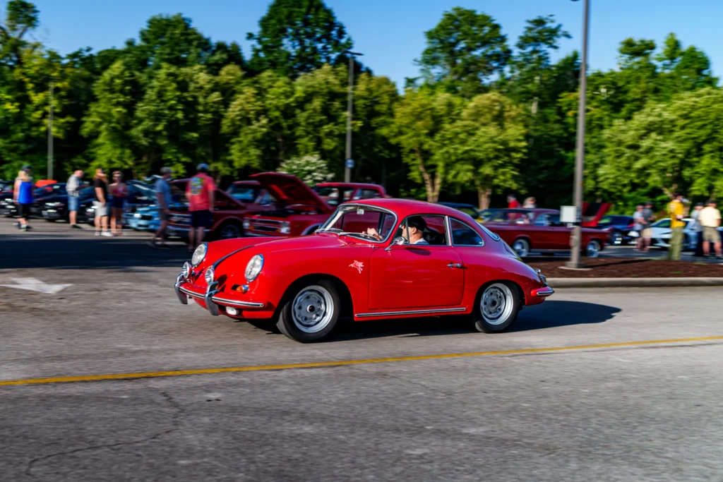 Red Porsche 356 driving past a parking lot with various cars and people