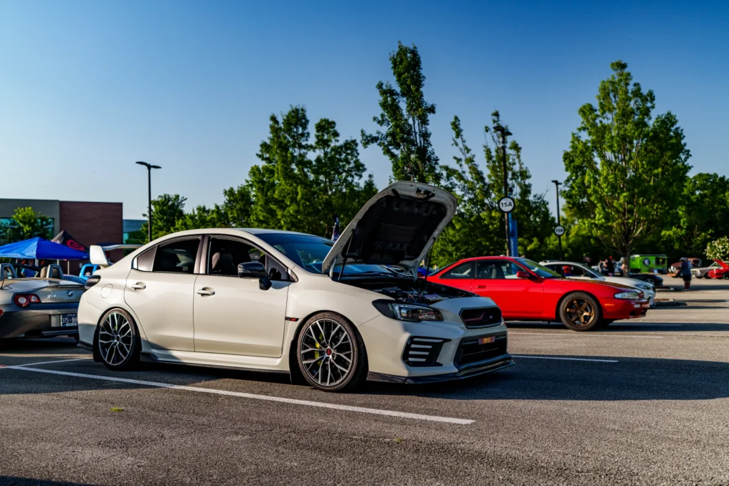 White Subaru WRX STI with hood open parked next to a red coupe and gray BMW Z4 at outdoor car event