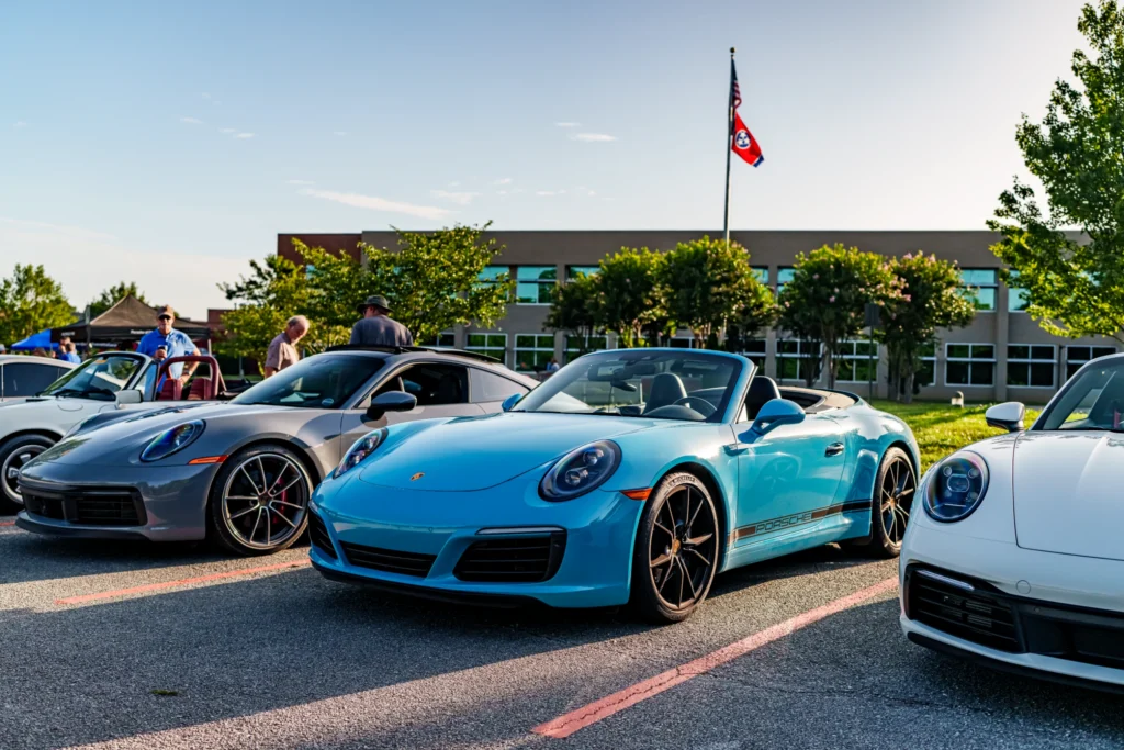 Three Porsche sports cars in parking lot at car meet, with people and building in background under clear sky.