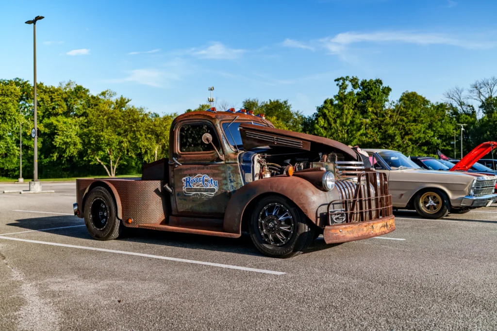 Rust-colored 46 Chevroletd truck with 'River City' sign and hood open in a parking lot with trees in the background