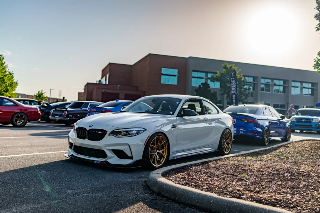 White BMW M2 with bronze wheels parked at an outdoor car meet with other cars and a building in the background