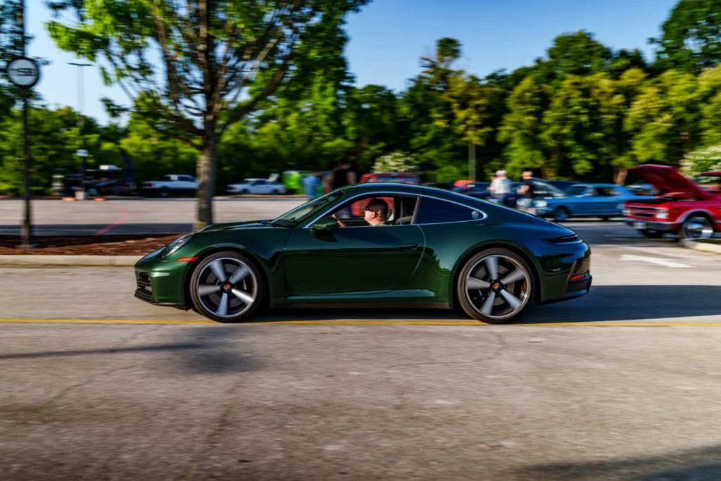 Dark green Porsche 911 coupe driving in parking lot with trees and classic cars in background
