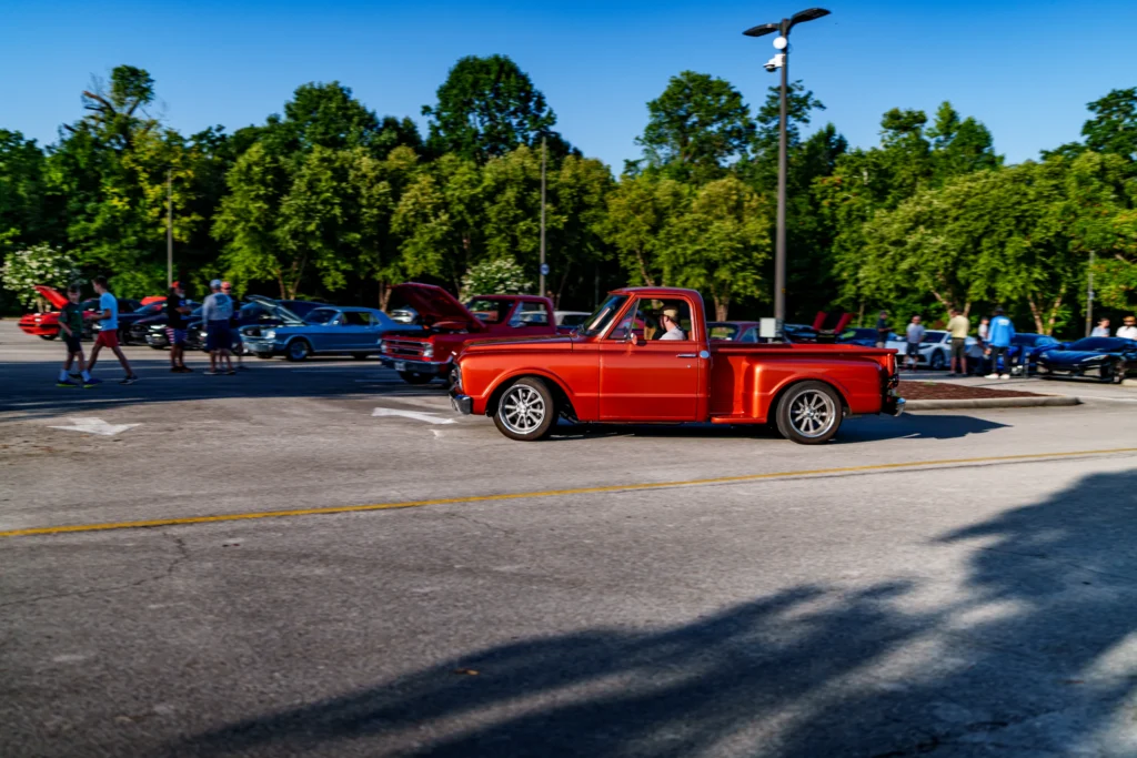 Orange vintage Chevrolet C10 pickup truck driving in a parking lot with other classic cars and attendees in the background