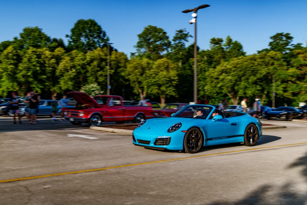 Blue Porsche 911 convertible driving in a parking lot at a car meet with a red vintage pickup and other cars in the backgroun