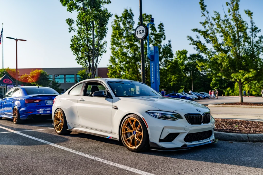 White BMW M2 with gold wheels parked next to a blue Audi sedan in a lot with trees and a building in the background