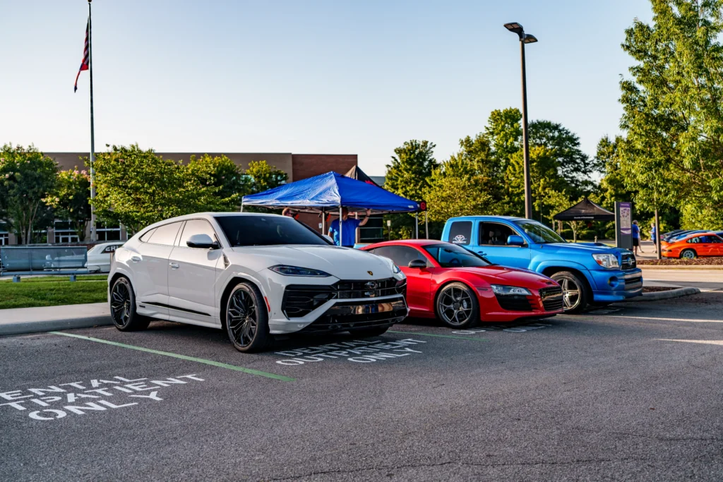 White Lamborghini Urus SUV and red Audi R8 sports car parked side by side at an outdoor car event in a parking lot