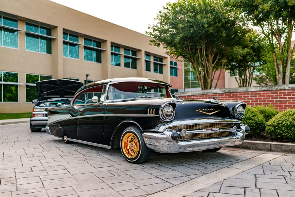 Black 1957 Chevrolet Bel Air with gold wheels parked in front of a building with other vehicles and trees nearby