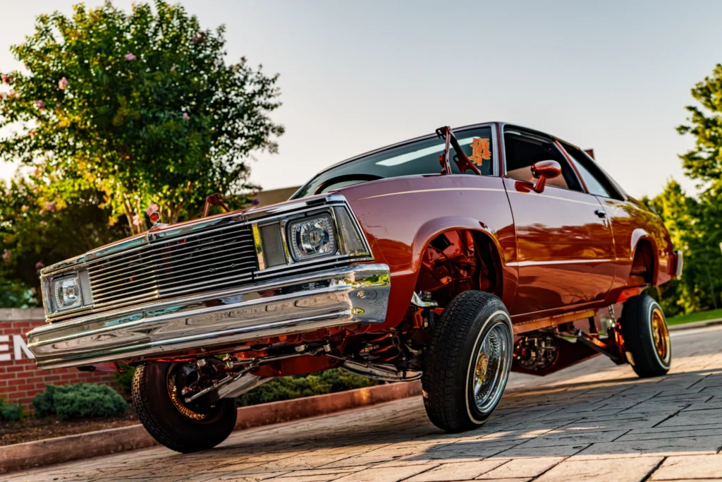 Reddish-brown lifted Chevrolet Malibu with hydraulic suspension in an outdoor setting with trees and brick wall