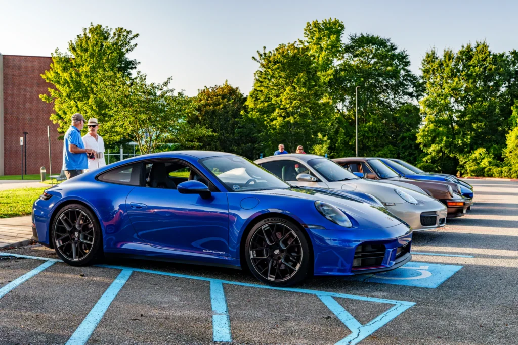 Four Porsche sports cars lined up in a parking lot with trees and people in the background.