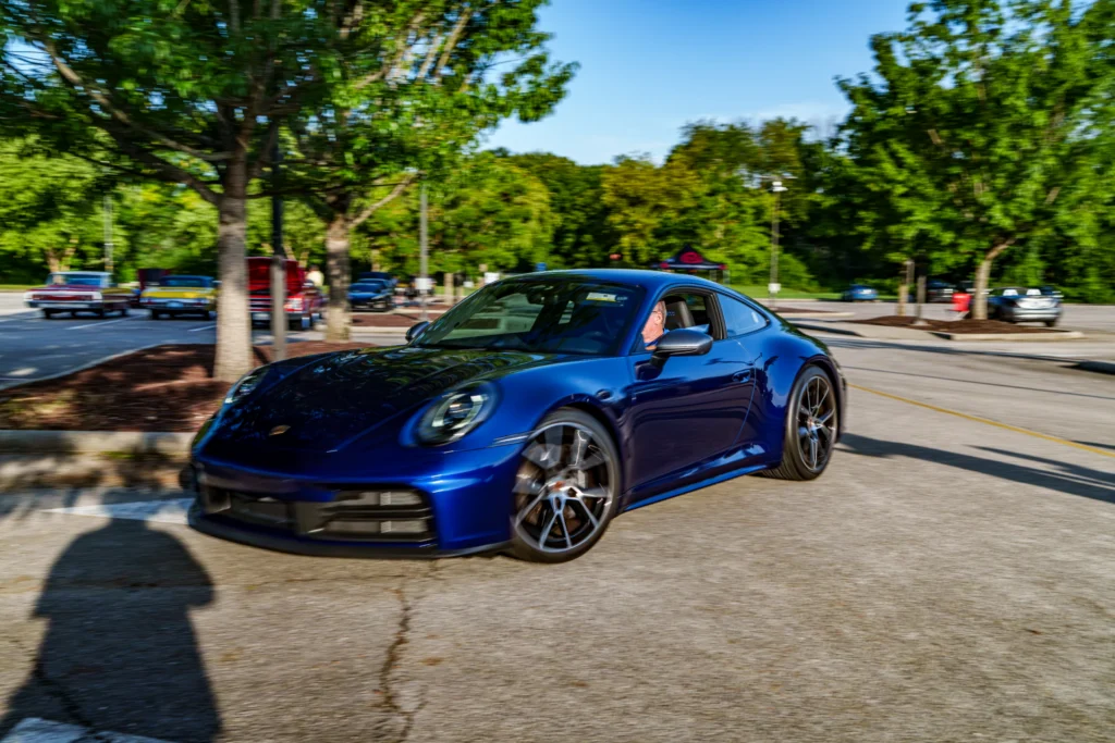 Blue Porsche 911 turning in a parking area with trees and other cars in the background