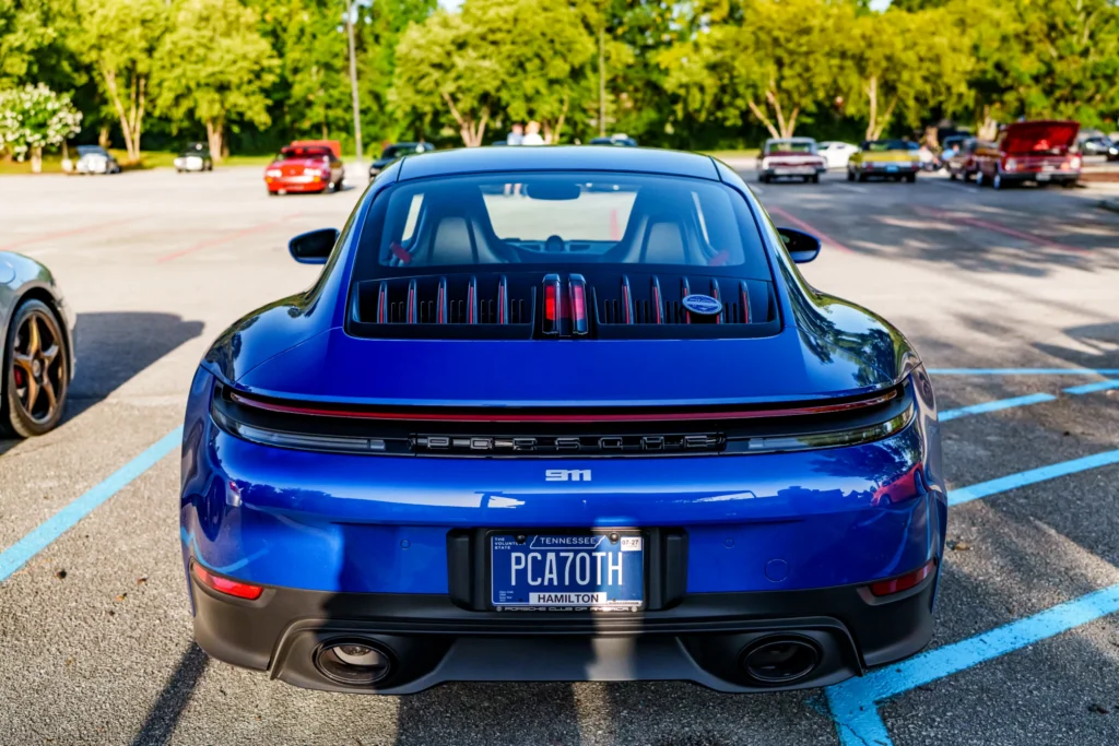 Rear view of blue Porsche 911 parked in lot with other classic and modern cars under trees