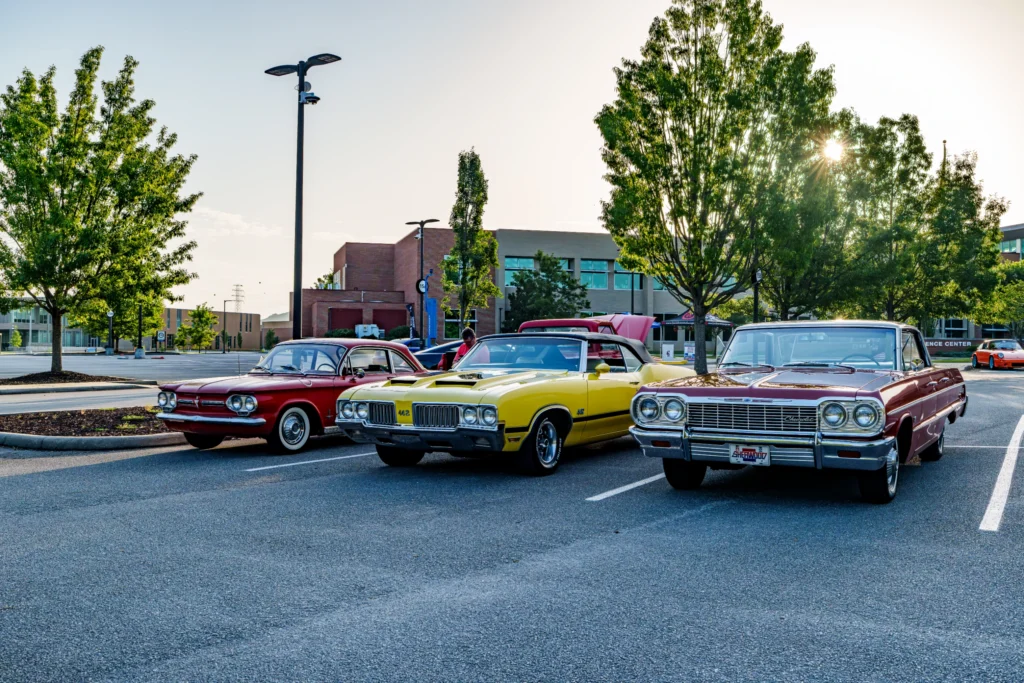 Red Chevrolet Corvair, yellow Oldsmobile 442 convertible, and maroon Chevrolet Impala parked in a lot with trees and building