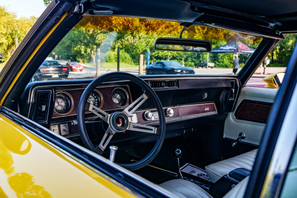 Interior view of a yellow Oldsmobile 442 with black steering wheel and white seats at an outdoor car event