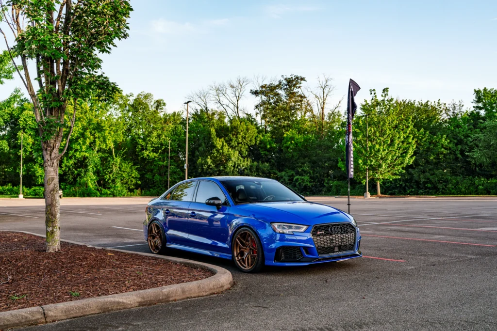 Blue Audi RS3 sedan parked in a mostly empty outdoor lot with a tree and flag nearby