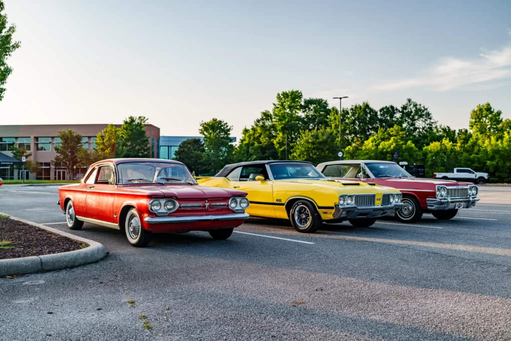 Red Chevrolet Corvair and yellow Oldsmobile 442 convertible parked next to a red Chevrolet Chevelle in a parking lot with tre