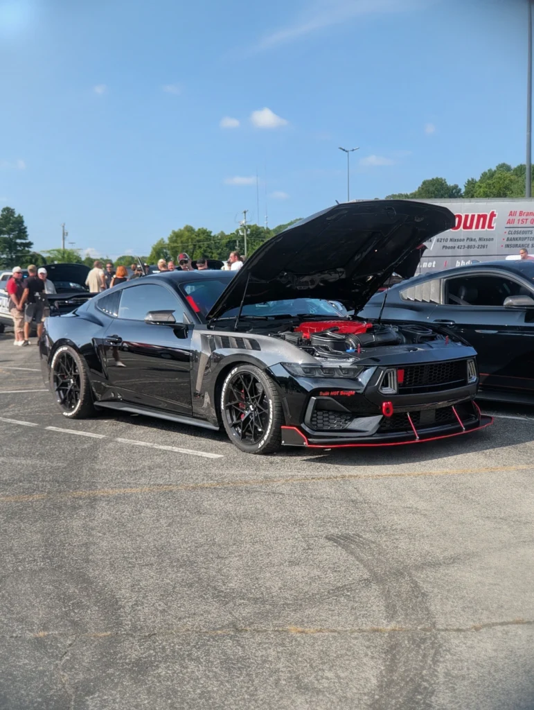 Black Ford Mustang with hood up showing engine, parked at a car event with people and other vehicles in background