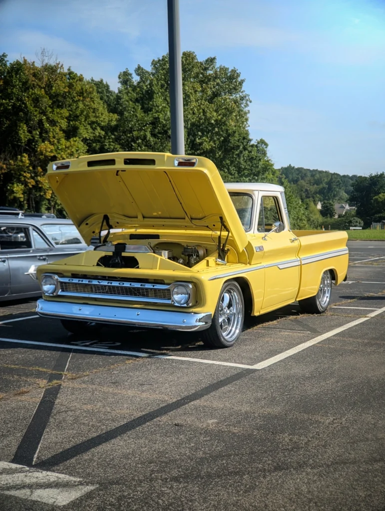 Yellow Chevrolet C10 pickup parked with its hood open in an outdoor lot on a clear day, with a silver car beside it and trees