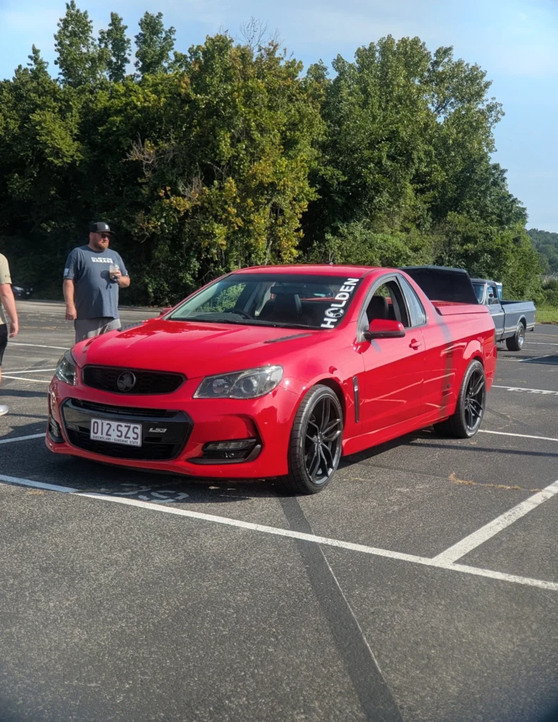Red Holden Ute in parking lot with trees in background and two men standing nearby