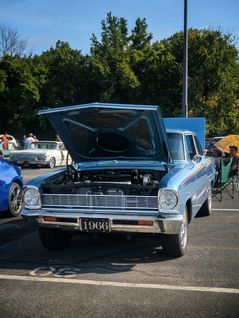Light blue 1966 Chevrolet Nova with hood open at outdoor car event on sunny day