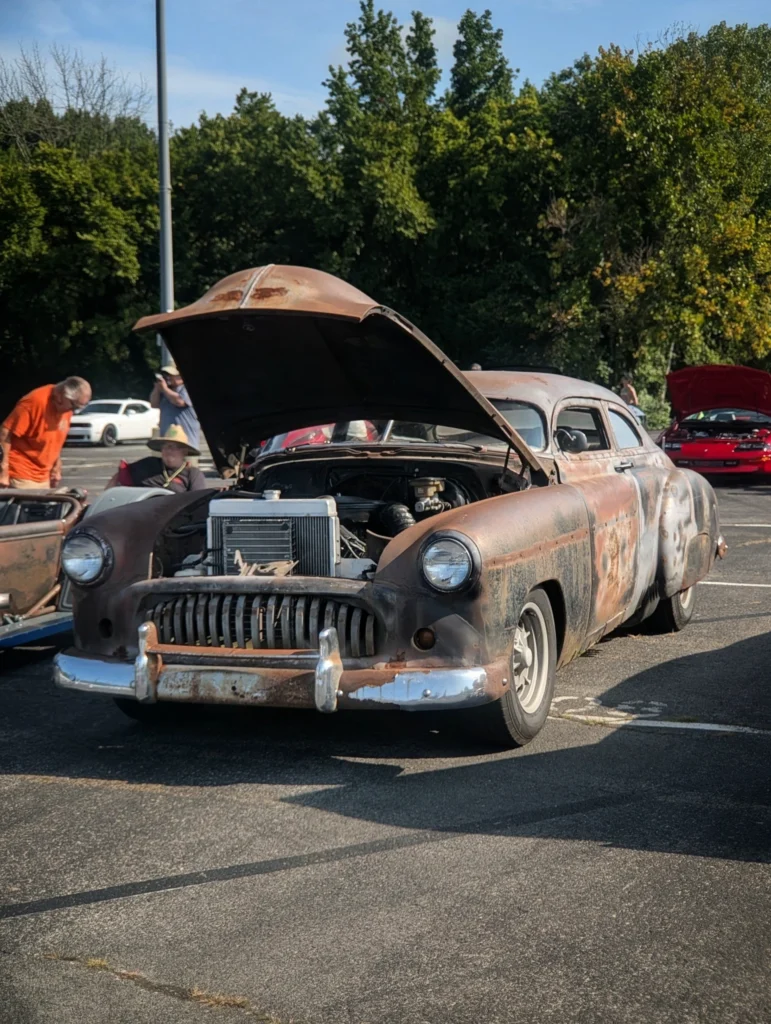 Rusty, weathered Buick Roadmaster with hood open at an outdoor car event with other vehicles and people in background