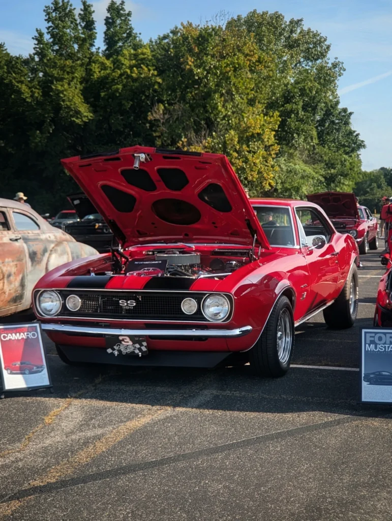 Red Chevrolet Camaro SS with hood open at an outdoor car event, surrounded by other vehicles and green trees in the backgroun