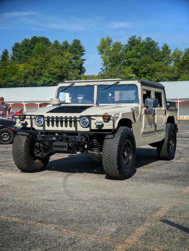 Beige Hummer H1 parked outdoors at a car event with trees and a building in the background