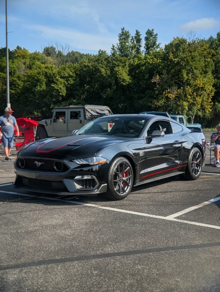 Black Ford Mustang Mach 1 parked in an outdoor lot with trees and people nearby