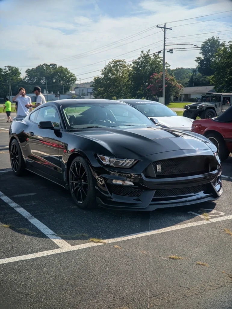 Black Ford Mustang Shelby GT350 parked in a lot with other vehicles and people in the background on a sunny day