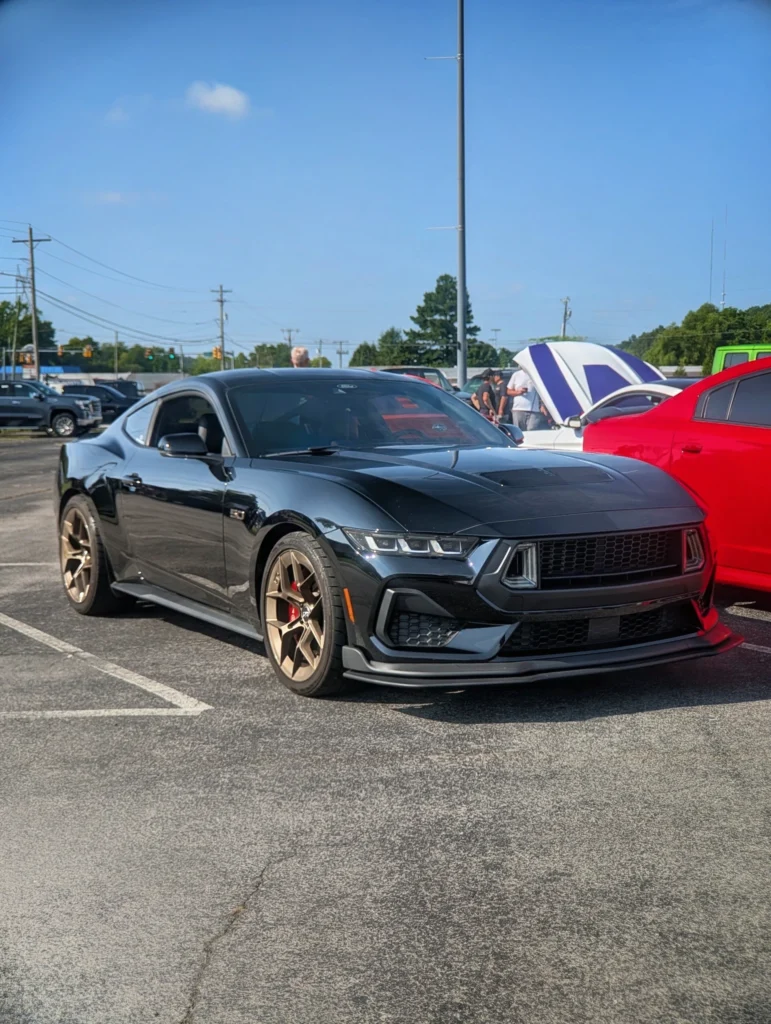 Black Ford Mustang GT with bronze wheels parked in a lot at a car event on a clear day