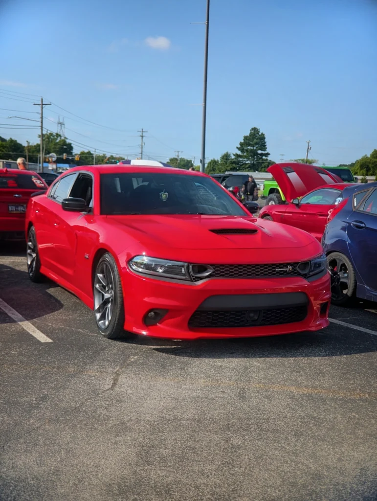 Red Dodge Charger parked in a lot at a car event with other vehicles and people in the background under clear sky.