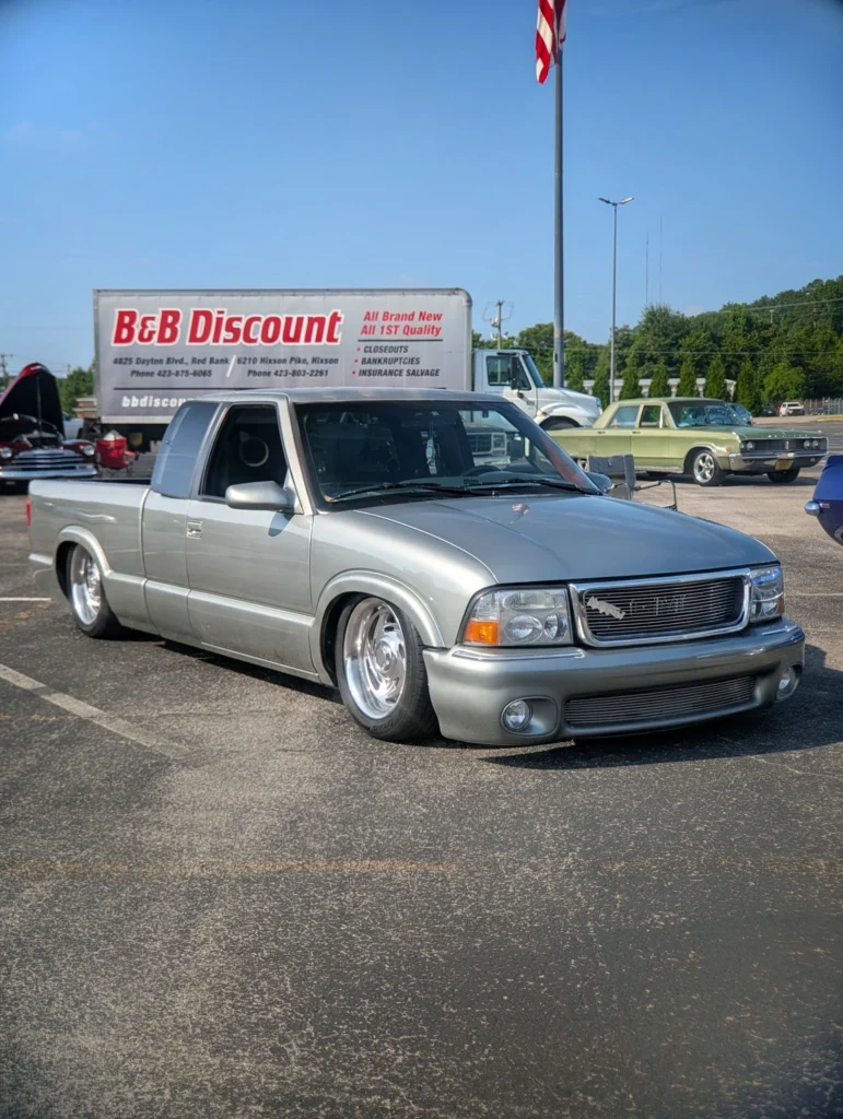 Silver GMC Sonoma pickup truck parked in a lot with a green sedan and a delivery truck in the background