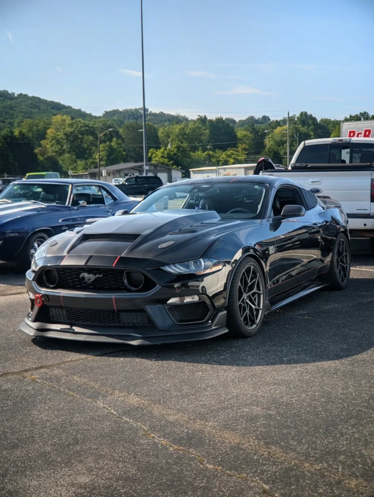 Black Ford Mustang with red accent stripes parked in a lot with trees and other cars in the background