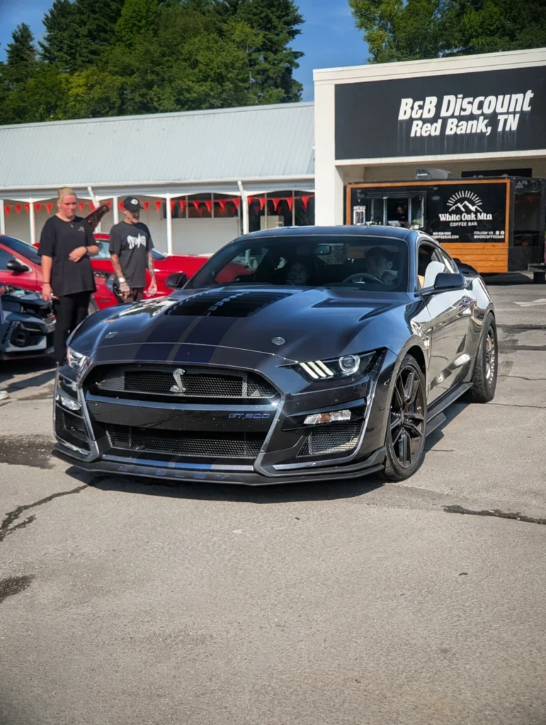 Black Ford Mustang Shelby GT500 with racing stripes in a parking lot near a discount store and coffee bar food truck