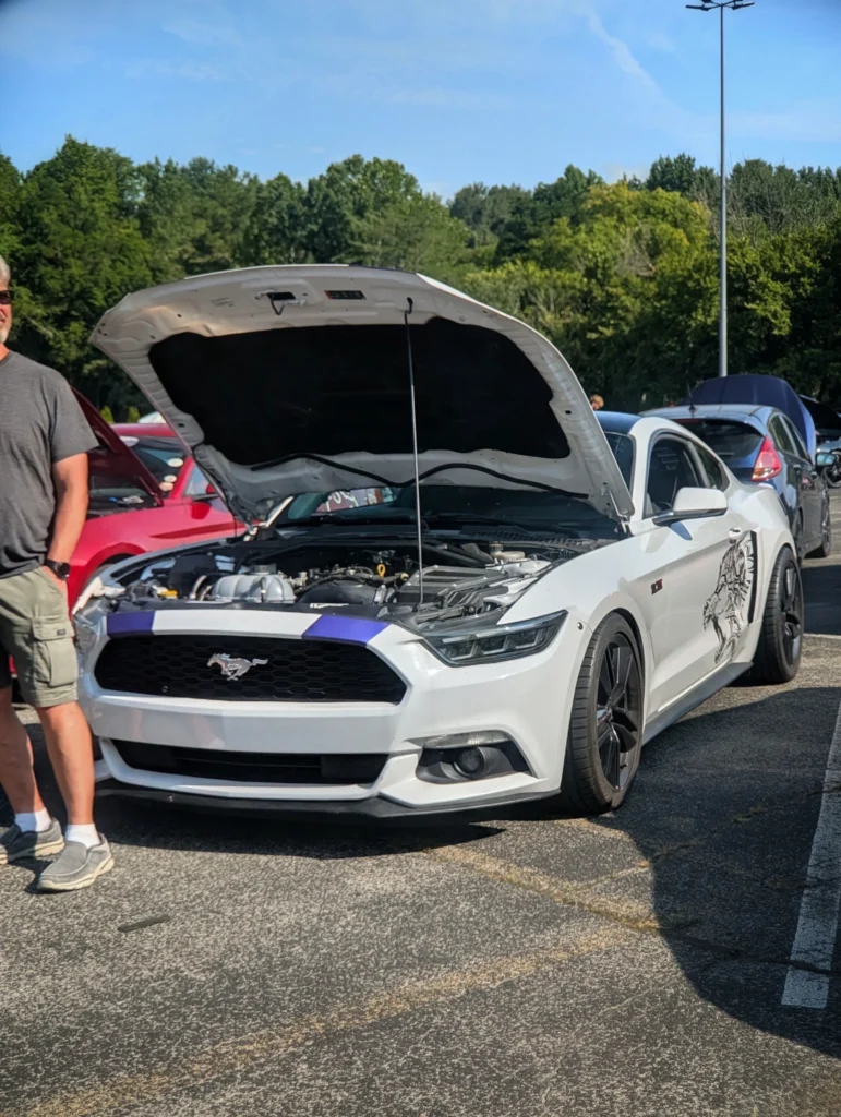 White Ford Mustang with hood open parked outdoors at a car event with trees and other cars in the background