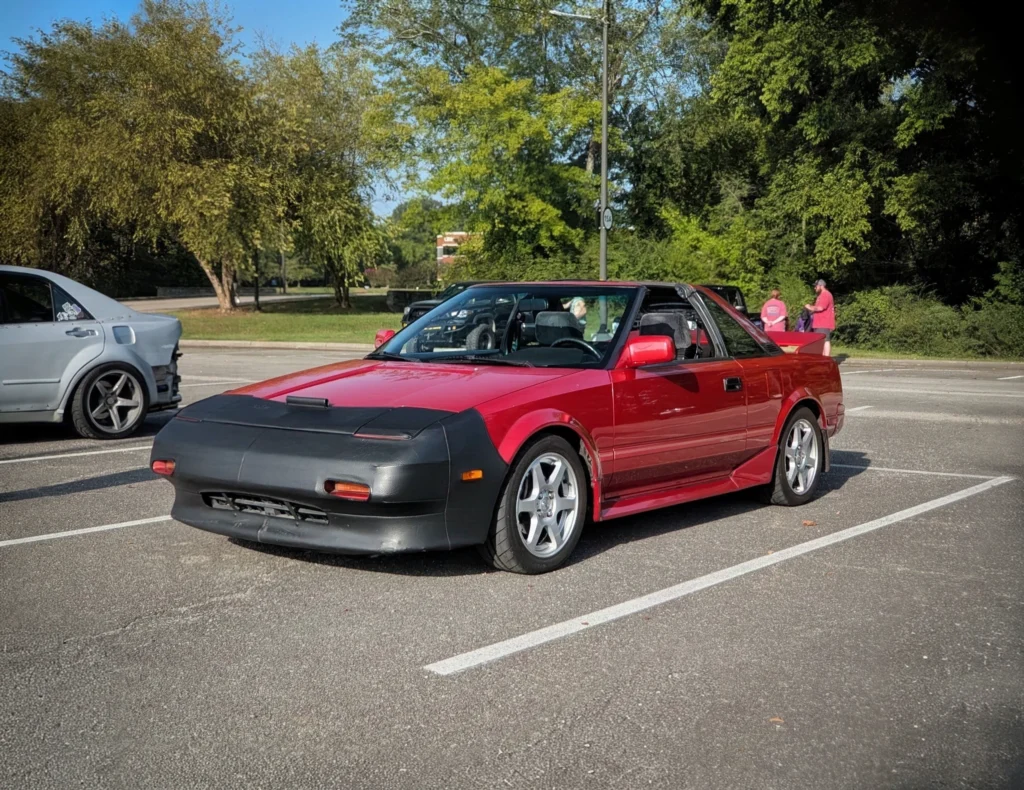 Red Toyota MR2 with black front bra parked beside a silver sedan in an outdoor parking lot with trees in the background