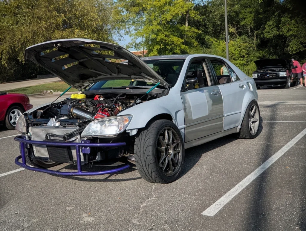 Silver Lexus IS sedan with hood open showing engine, parked in a lot with trees and other vehicles in background