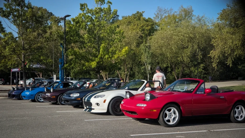 A lineup of five Mazda Miatas in various colors parked in an outdoor lot with trees in the background