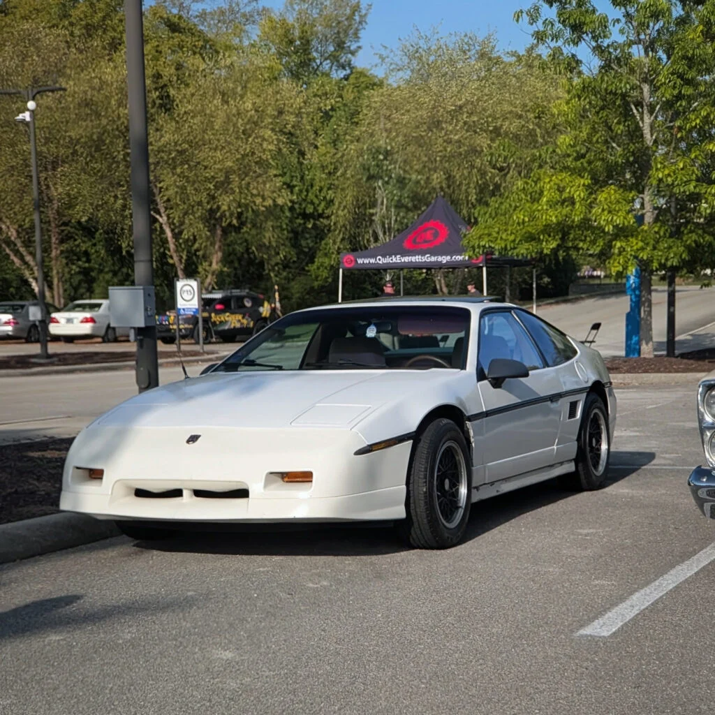 White Pontiac Fiero GT parked in an outdoor lot with trees and a QuickEverettsGarage tent in the background