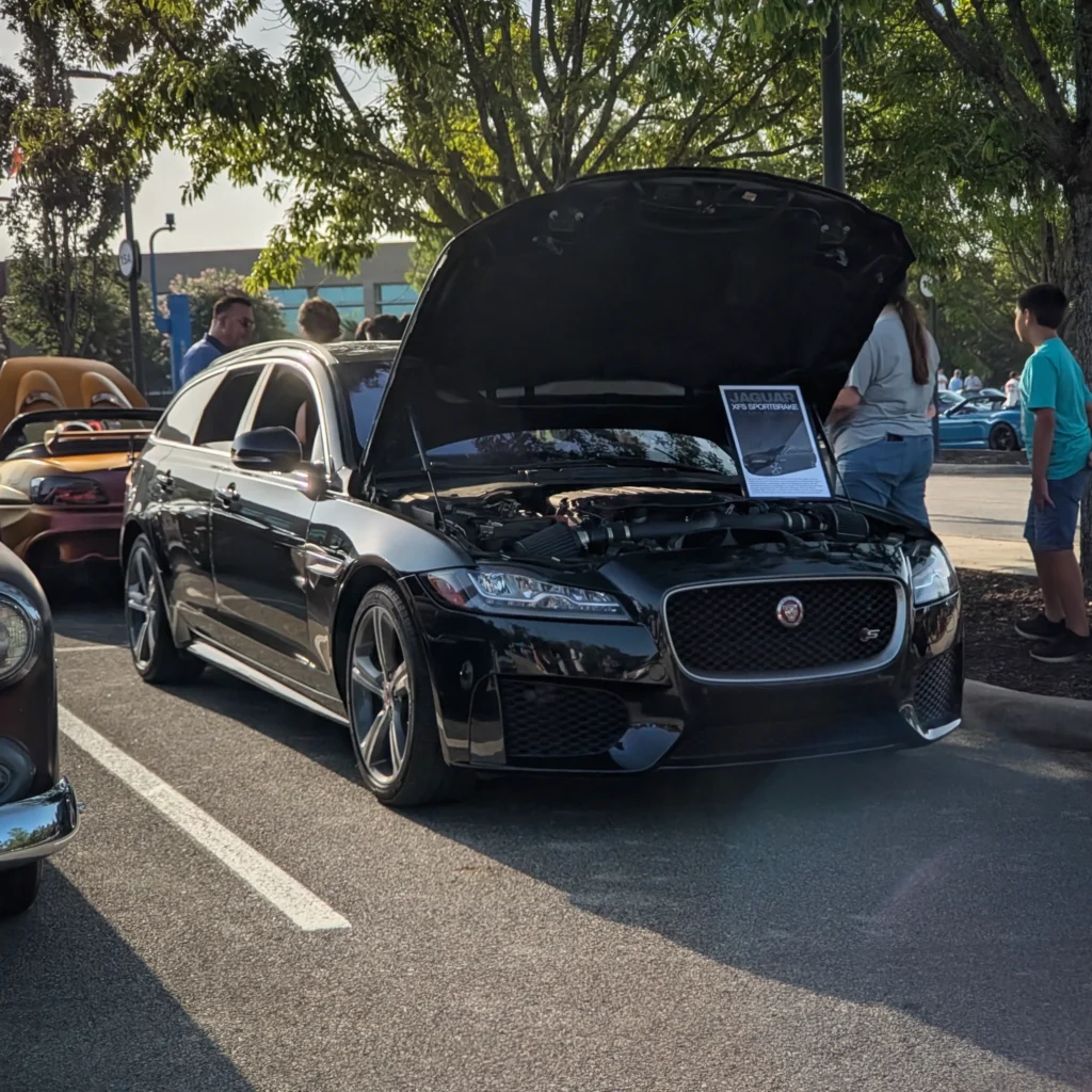 Black Jaguar XFS Sportbrake with open hood at outdoor car event with trees and people nearby