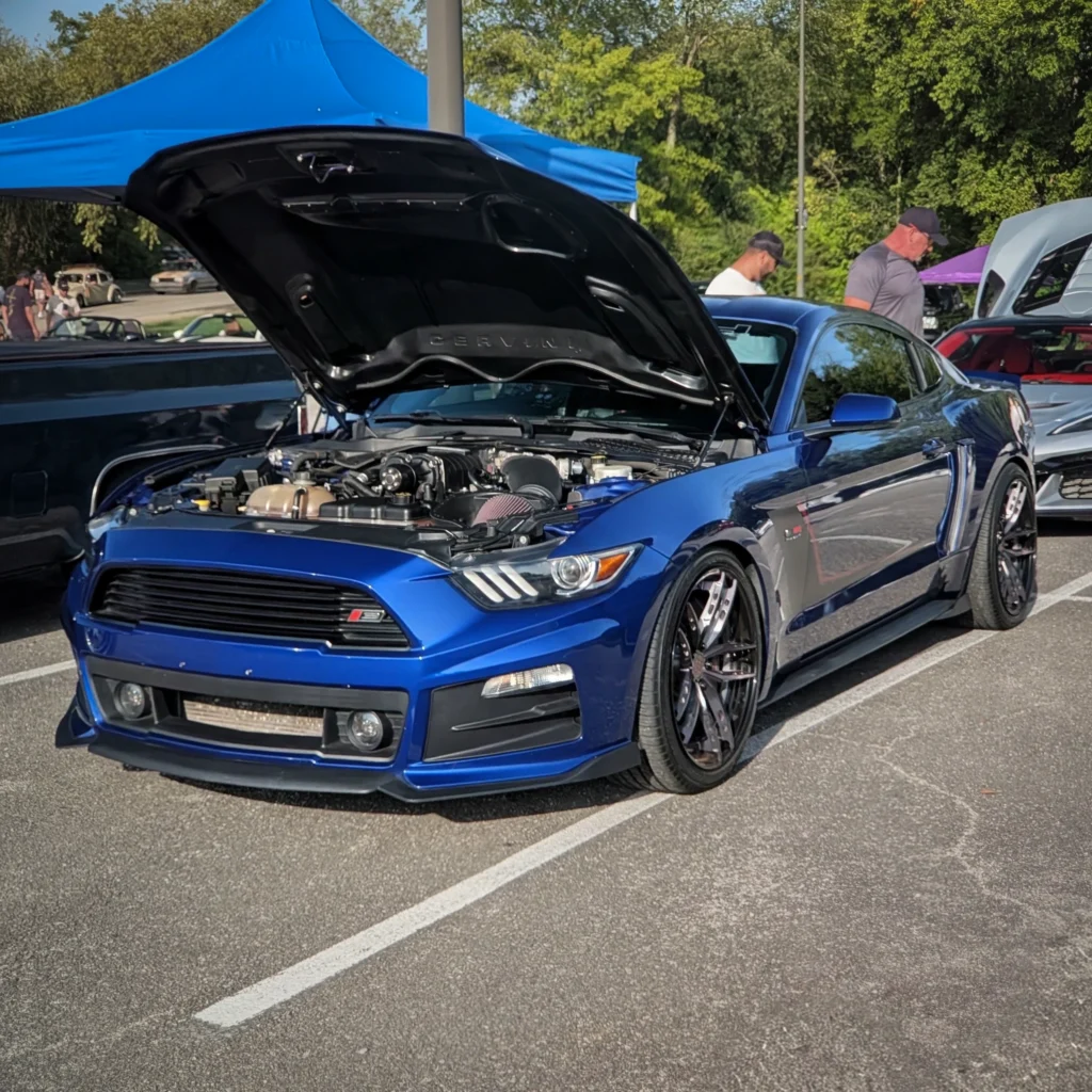 Blue Ford Mustang GT with hood open at car meet in sunny parking lot