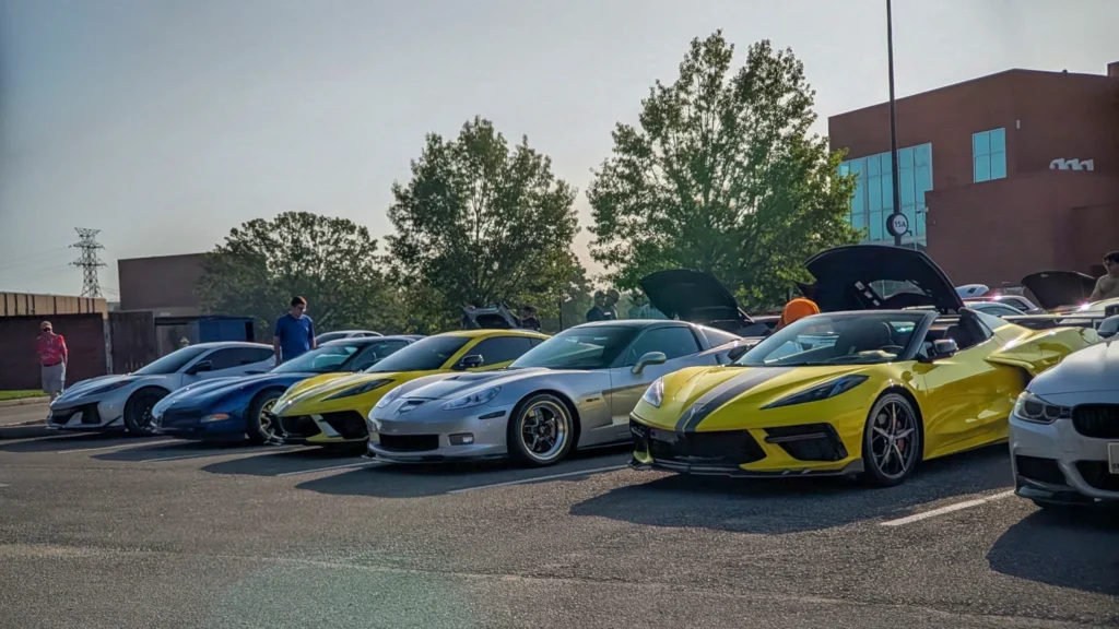 Lineup of sports cars including yellow and silver Chevrolet Corvettes in a parking lot at a car meet during daytime