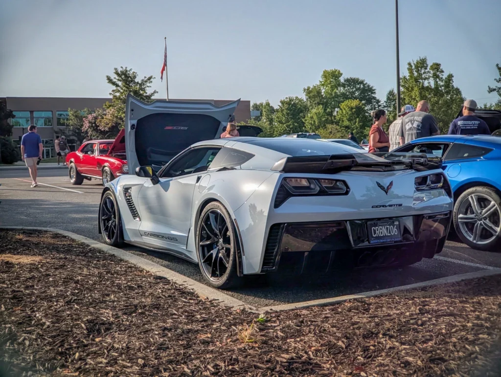 White Chevrolet Corvette Z06 with rear hatch open at an outdoor car event parking lot with people and other cars nearby.