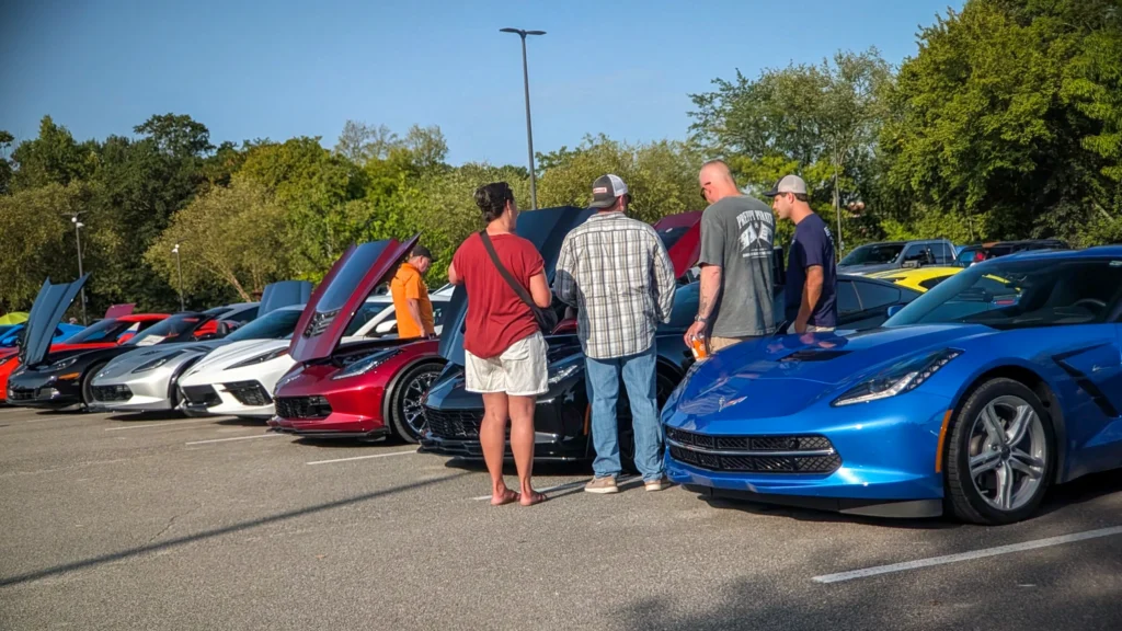 A lineup of Corvettes in various colors at an outdoor car meet with several people standing and looking at cars with open hoo