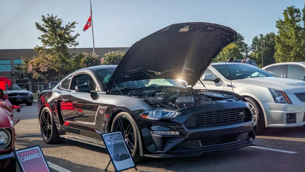 Black Ford Mustang with hood open in a parking lot during daytime car event
