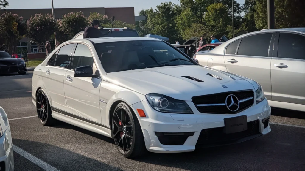 White Mercedes-Benz C63 AMG sedan parked at an outdoor car event with a black car and people in the background