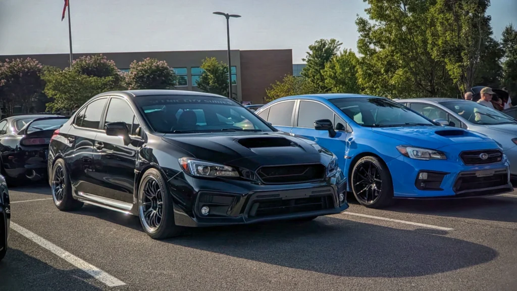 Black and blue Subaru WRX sedans parked side by side at an outdoor car event in a parking lot with trees and a building in th