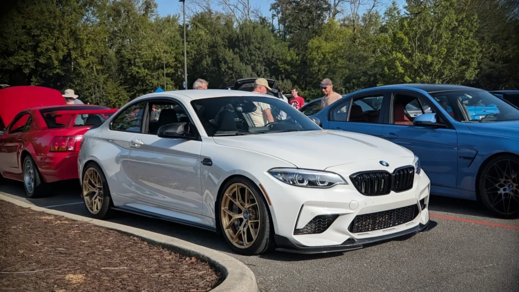White BMW M2 with bronze wheels parked with a red coupe and a blue sedan at an outdoor car event with people nearby
