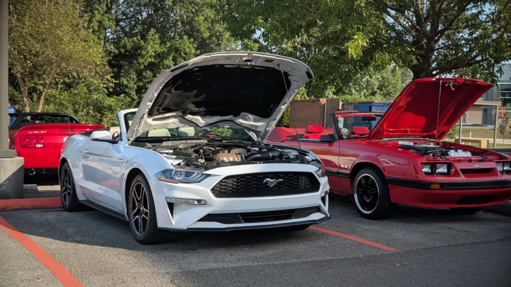 White Ford Mustang convertible and red Ford Mustang convertible with hoods open parked outdoors with trees in background